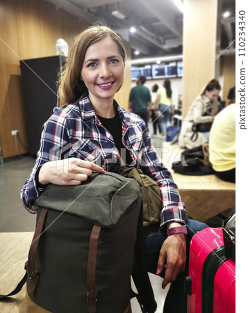 Young woman in shirt waiting for her transfer, with her backpack and luggage near her, looking into camera, smiling, blurred station in background Young woman in shirt waiting for her transfer, with her backpack and luggage near her, looking into camera, smiling, blurred station in background 110234340