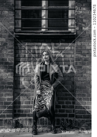 Stylish blond woman posing near red brick wall. Beautiful fashionable girl stands near the brick wall. Fashionable woman in spectacles hanging out in night city. 110234678