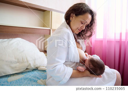 Portrait of a mother and her cute newborn baby. Happy mom smiles looking at her baby boy while breastfeeding him 110235005