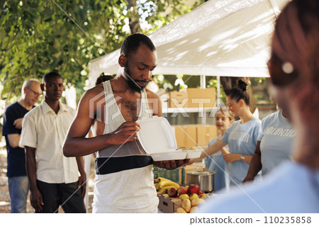 Image of african american homeless guy holding his free food provided by the nonprofit group. Volunteers working to feed the needy, underprivileged and disadvantaged at an outdoor food bank. 110235858