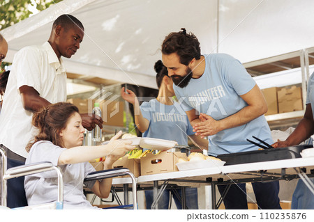 Friendly male volunteer assists and serves free food to the poor needy woman in wheelchair. Charity workers at an outdoor food bank providing hunger relief to the disabled and less fortunate. 110235876