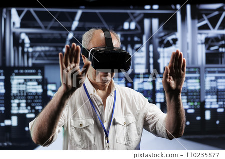 Elderly adept technician immersed in virtual reality at data center, doing infrastructure maintenance. Specialist using VR headset to optimize server clusters system performance 110235877