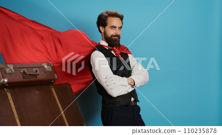 Hotel worker with red mantle to help guests with trolley bags, pretending to be fantasy cartoon superhero character on camera. Bellhop acting like comic superhuman with cape against background. Hotel worker with red mantle to help guests with trolley bags, pretending to be fantasy cartoon superhero character on camera. Bellhop acting like comic superhuman with cape against background. 110235878