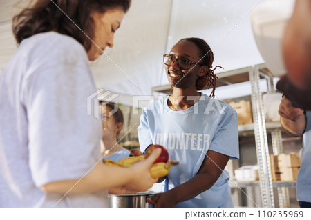 Portrait of female charity worker handing out fresh fruits and food to poor and homeless people. Cheerful black woman volunteers at food drive serving free meals to the hungry and less fortunate. Portrait of female charity worker handing out fresh fruits and food to poor and homeless people. Cheerful black woman volunteers at food drive serving free meals to the hungry and less fortunate. 110235969