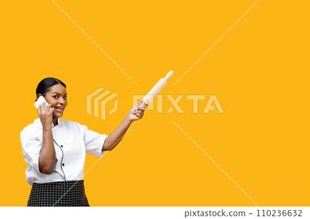 Efficient black female chef managing tasks over phone and holding rolling pin Efficient black female chef managing tasks over phone and holding rolling pin 110236632