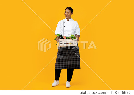 Smiling black female chef presenting wooden crate of organic vegetables 110236652
