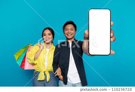 Happy Asian couple with colorful shopping bags and a modern smartphone, presenting a blank phone screen 110236728