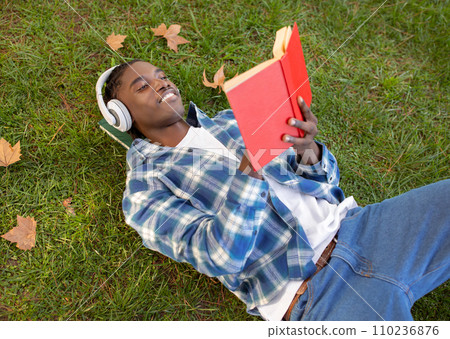 Relaxed black young man enjoying book with headphones lying outside Relaxed black young man enjoying book with headphones lying outside 110236876