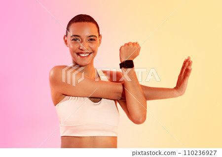 Radiant young woman with a joyful smile stretches her arm muscles while wearing a fitness tracker 110236927