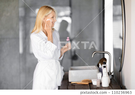 Smiling mature woman wearing white bathrobe applying refreshing facial toner in bathroom Smiling mature woman wearing white bathrobe applying refreshing facial toner in bathroom 110236963