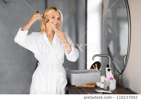 Smiling senior woman using green jade facial roller for her skincare routine Smiling senior woman using green jade facial roller for her skincare routine 110237002