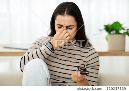 A young woman with a pained expression pinches her nose and squints, holding eyeglasses 110237189