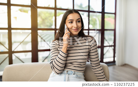 Smiling millennial caucasian lady call by phone at sofa, talk in living room interior Smiling millennial caucasian lady call by phone at sofa, talk in living room interior 110237225