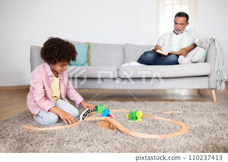 grandson playing building toy railway while senior grandpa reading indoors grandson playing building toy railway while senior grandpa reading indoors 110237413