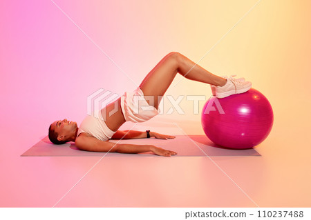 A woman with a shaved head lies on a yoga mat doing a hamstring curl on a pink exercise ball A woman with a shaved head lies on a yoga mat doing a hamstring curl on a pink exercise ball 110237488