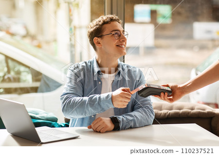 Cheerful young european man using credit card for payment machine, sit on desk with laptop 110237521