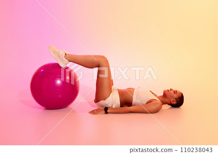 A relaxed woman with a shaved head lies on her back performing a bridge exercise with her feet 110238043