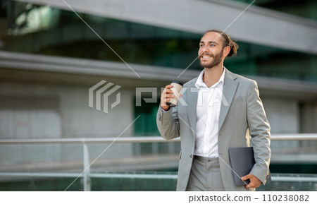 Relaxed young businessman holding laptop and cup of coffee while walking outdoors 110238082