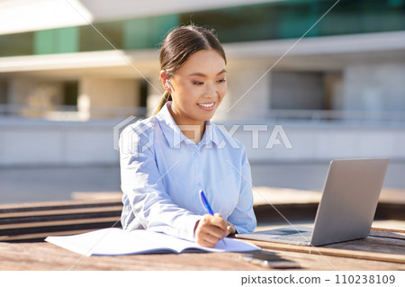 Korean business lady near laptop browsing and writing, sitting outdoors Korean business lady near laptop browsing and writing, sitting outdoors 110238109