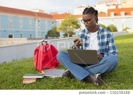 African university student guy with laptop scrolling phone sitting outside 110238188