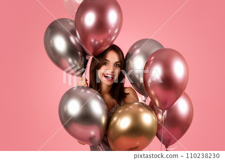 A delighted woman with dark hair and red lipstick smiles joyfully, surrounded by a cluster of metallic balloons 110238230