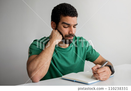 Serious man with a beard in a green t-shirt writing in a notebook, resting his chin 110238331