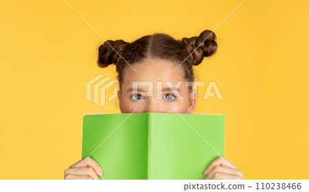 Curious young girl with buns hairstyle peeks over a green book, her eyes wide with intrigue Curious young girl with buns hairstyle peeks over a green book, her eyes wide with intrigue 110238466