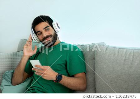 Relaxed young man with a beard and a green shirt, lounging on a gray couch 110238473