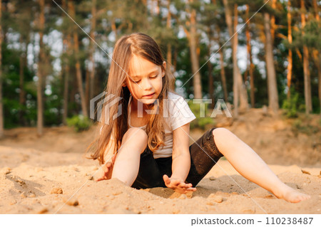 Little girl with dark hair sit on sand near forest. Female child in white t-shirt and black shorts have fun in nature. 110238487