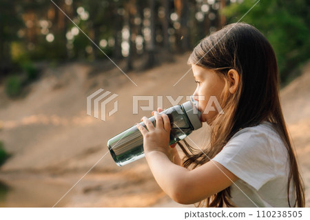 Little girl with dark hair drinking water from bottle sitting outdoor. Active kid resting on sandy beach in nature. 110238505