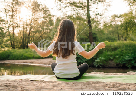 Little girl sit on sporty mat on plage in yoga pose with arms wide open. Kid with dark hair meditating next to lake. Little girl sit on sporty mat on plage in yoga pose with arms wide open. Kid with dark hair meditating next to lake. 110238526