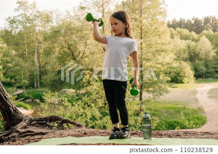 Dynamic little sportsman working out with dumbbells in hands on sporty mat outdoor. Girl lifting weights in nature. Dynamic little sportsman working out with dumbbells in hands on sporty mat outdoor. Girl lifting weights in nature. 110238529