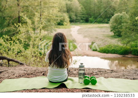 Little female sportsman sit on mat on hill in front of lake and enjoy nature. Dumbbells and bottle lying near child. Little female sportsman sit on mat on hill in front of lake and enjoy nature. Dumbbells and bottle lying near child. 110238533