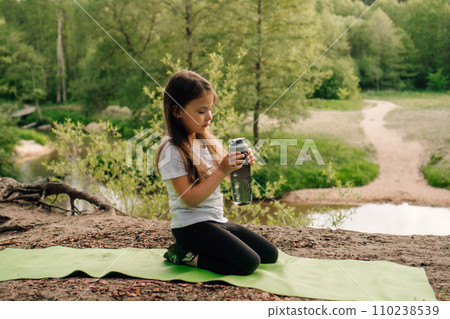 Little brunette lady calmly sit on sporty mat outdoor and hold bottle. Child look at flask resting in front of river. 110238539