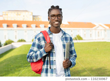 Friendly black student guy with backpack holding closed laptop outside 110238967