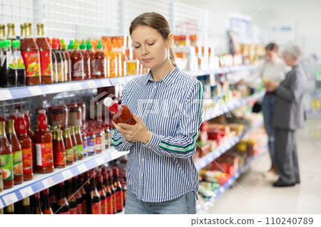 Woman customer is thinking about buying spicy and unusual red sriracha sauce. 110240789