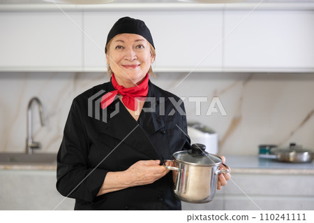 Elderly woman cook in chef hat and black uniform stands in kitchen and holds stainless pan Elderly woman cook in chef hat and black uniform stands in kitchen and holds stainless pan 110241111