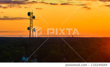 Cell tower above green forest glowing orange at sunset aerial with yellow sky 110244844