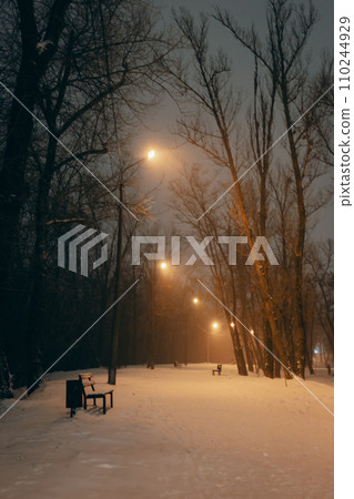Empty winter park with street lamps at night. Winter landscape. Snowfall in the city. Bench on the alley in evening park. Frozen weather background. February weather. Snow and freeze. Empty winter park with street lamps at night. Winter landscape. Snowfall in the city. Bench on the alley in evening park. Frozen weather background. February weather. Snow and freeze. 110244929