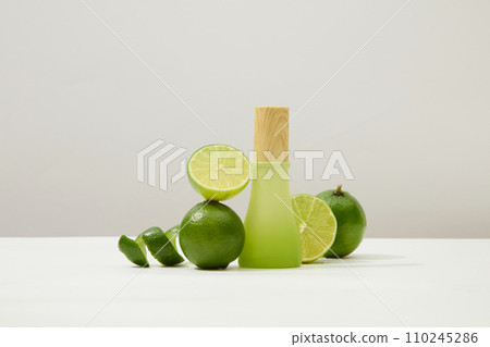 Front view of a green glass bottle with wooden lid, next to fresh lemon on a minimalist white background. Vitamin C in lemon helps restore damaged skin very quickly. 110245286