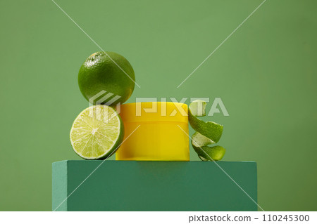 An unlabeled yellow cosmetic jar and fresh lemons are displayed on a podium with a green background. Lemon extract is effective in treating blackheads. 110245300