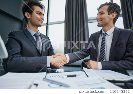 Smiling businessman shaking client hand, closing successful deal, sitting at table in office Smiling businessman shaking client hand, closing successful deal, sitting at table in office 110245352