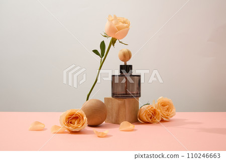 Front view of an essential oil bottle on a wooden platform, a wooden sphere and fresh roses standing out against a pastel background. Roses have a pleasant fragrance and are commonly used. Front view of an essential oil bottle on a wooden platform, a wooden sphere and fresh roses standing out against a pastel background. Roses have a pleasant fragrance and are commonly used. 110246663