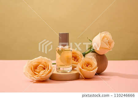 A labelless transparent perfume bottle is placed on a wooden platform with fresh roses. Natural cosmetics concept for advertising. Pink surface with brown background. 110246666