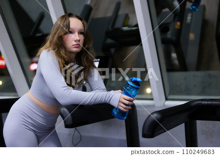Portrait of sports woman with water. beautiful strong muscular woman drinking water in gym. young woman in grey sportswear with bottle of water. Fit thin gimnast sitting on stairs 110249683