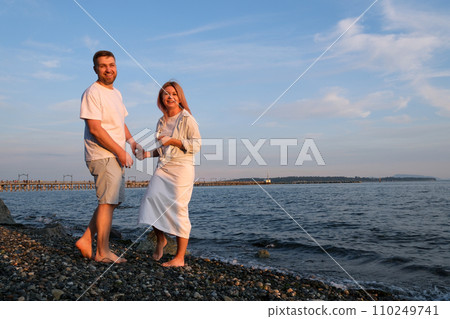 Couple spending time on the beach. Walking barefoot. Man wearing trousers and shirt. Woman wearing white dress. Relation between husband and wife. Romantic concept. Traveling couple. Copy space. Bali 110249741