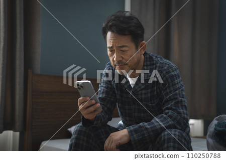 Middle-aged man sitting on bed and staring at smartphone screen 110250788