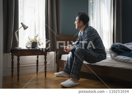 Middle-aged man in pajamas sitting on bed looking out the window 110251034
