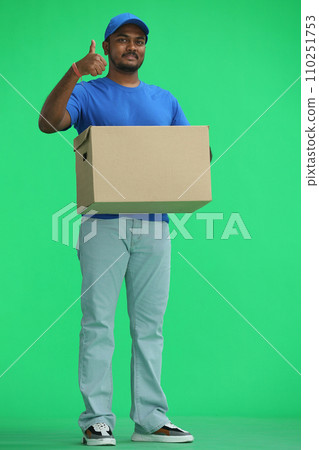 A male deliveryman with a box, on a green background, in full height, shows a thumbs up A male deliveryman with a box, on a green background, in full height, shows a thumbs up 110251753