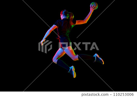 Fit young woman engaged in intense handball training, perfecting her throwing and catching abilities against black background in neon light, filter. Fit young woman engaged in intense handball training, perfecting her throwing and catching abilities against black background in neon light, filter. 110253006